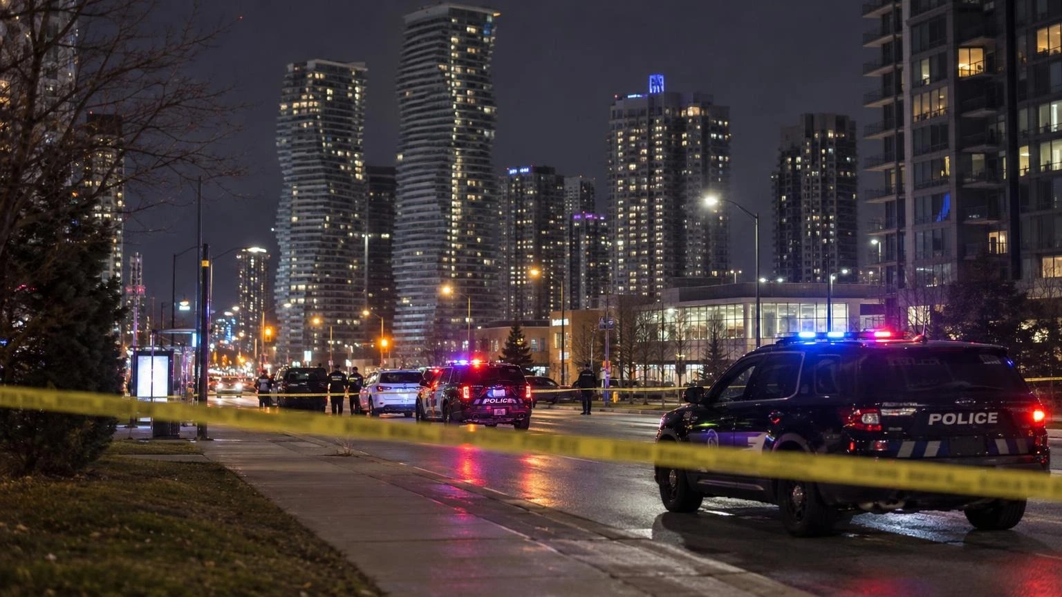 A close-up of yellow police tape stretched across a sidewalk at dusk, with blurred blue and red emergency lights in the background.