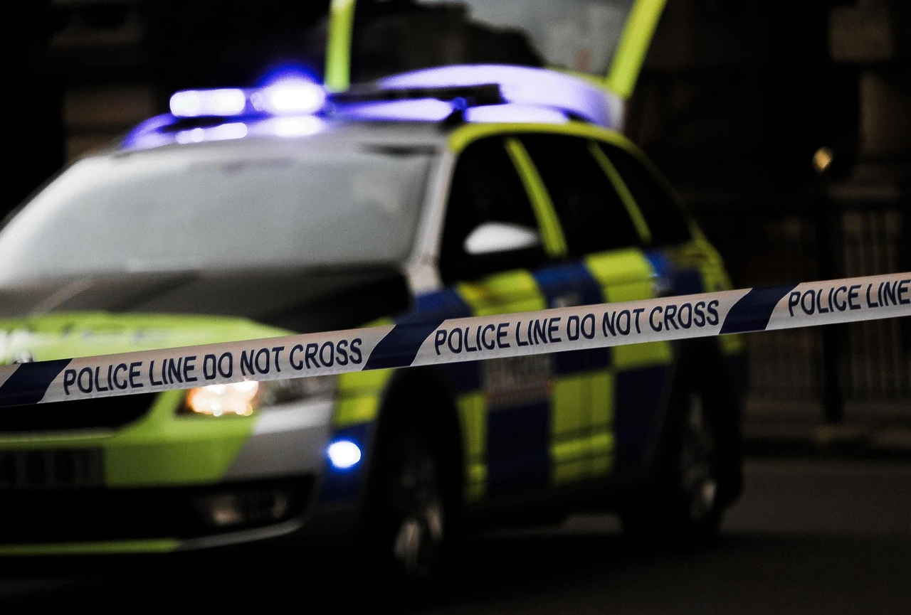 A close-up of yellow police tape stretched across a suburban residential area at dusk, with blurred emergency lights in the background.
