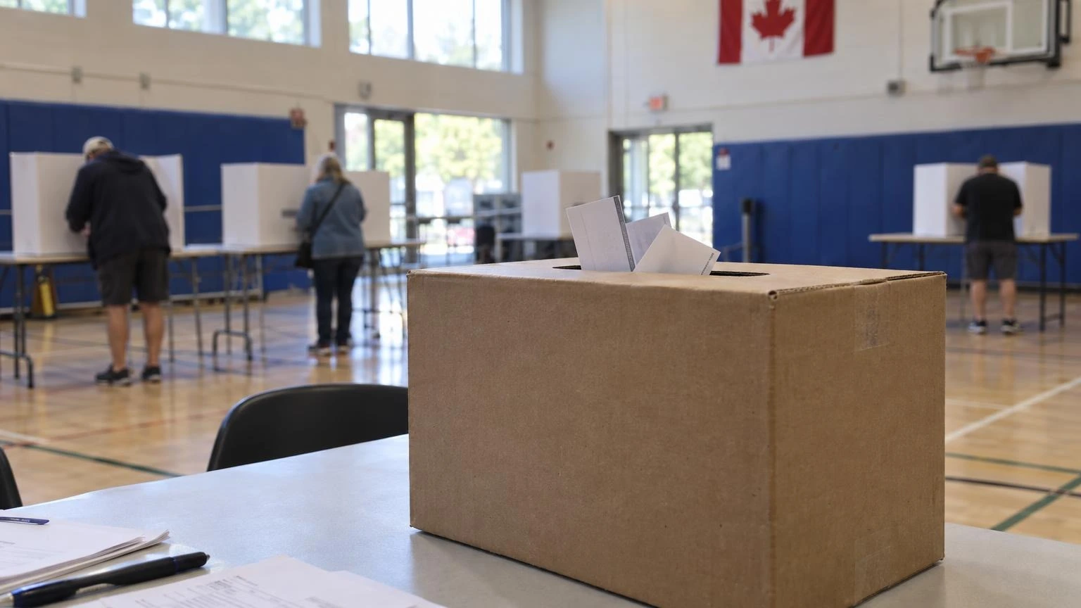 A close-up of several folded paper ballots inside a plain cardboard ballot box at a community centre.