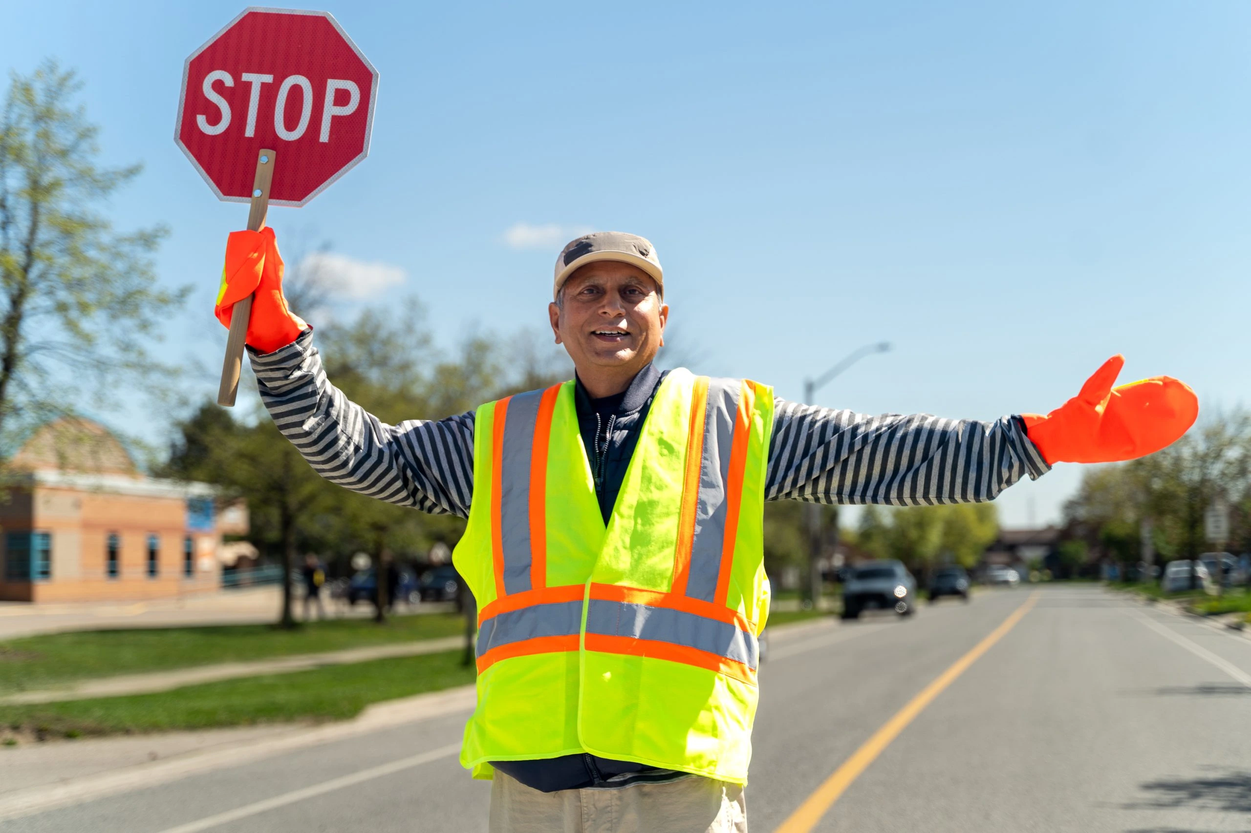 A crossing guard in a reflective vest holds a stop sign while children wait at a curb in a suburban school zone on a September morning.