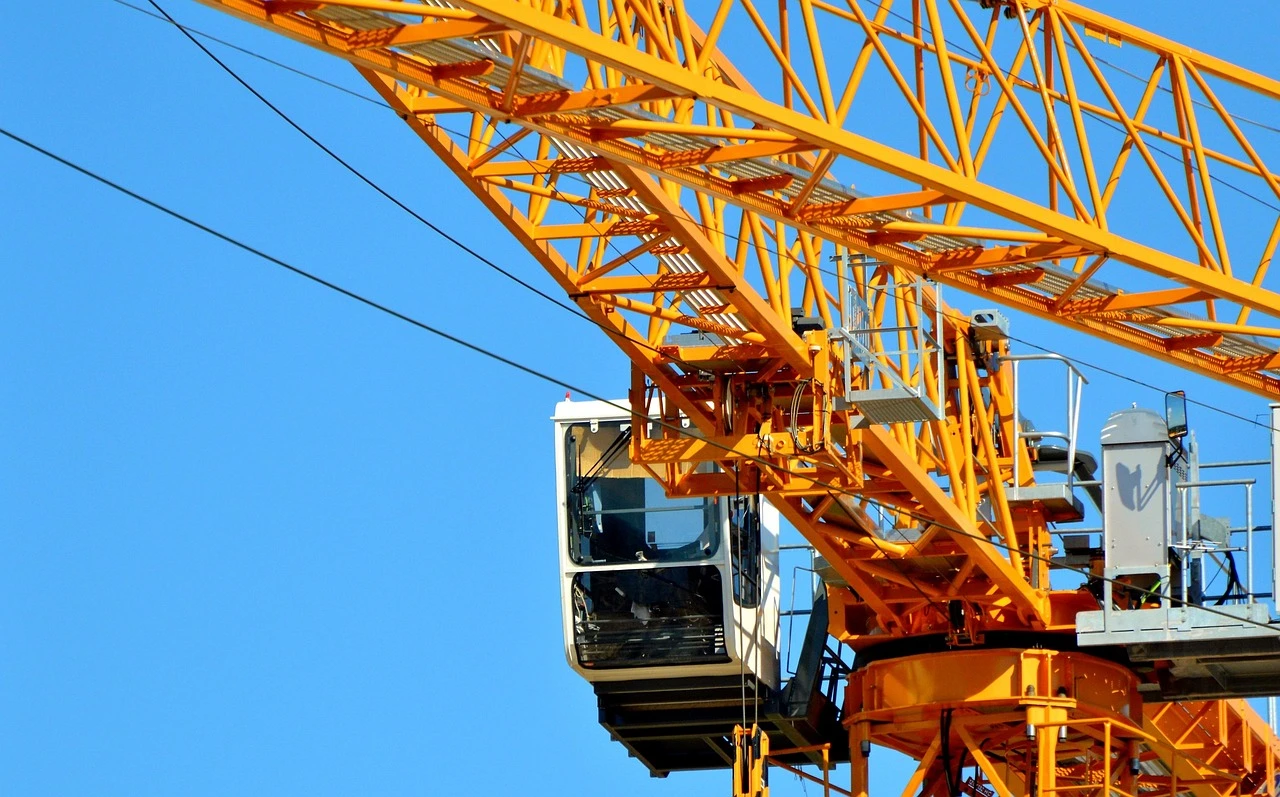 A hospital building under construction with tower cranes on an early-autumn day, framed by amber and bare trees.