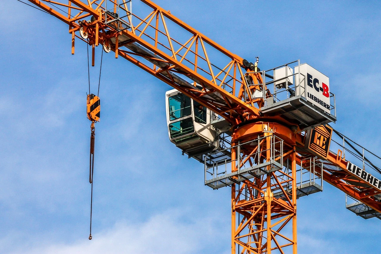A construction site with a crane and excavator on an urban lot beside brick commercial buildings under green summer trees.