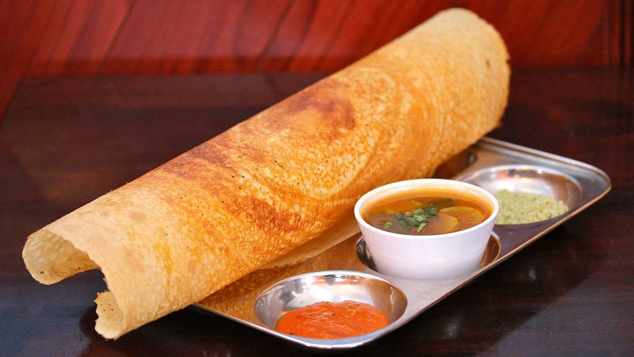 A plate with a rolled masala dosa and small bowls of sambar and chutneys on a restaurant table.