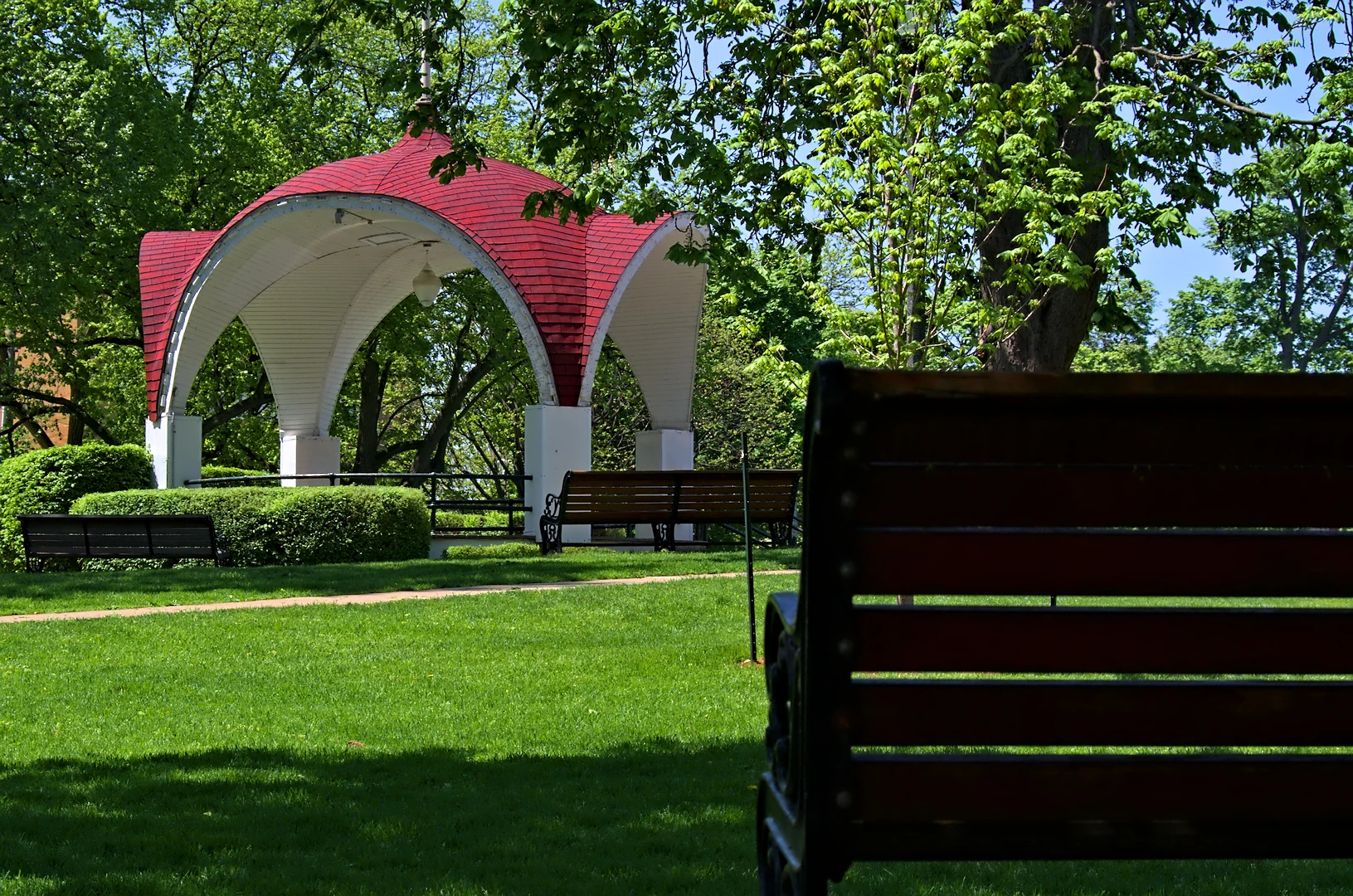 A green city park with a bandshell, trees strung with festival lights and people seated on the grass on a summer afternoon.