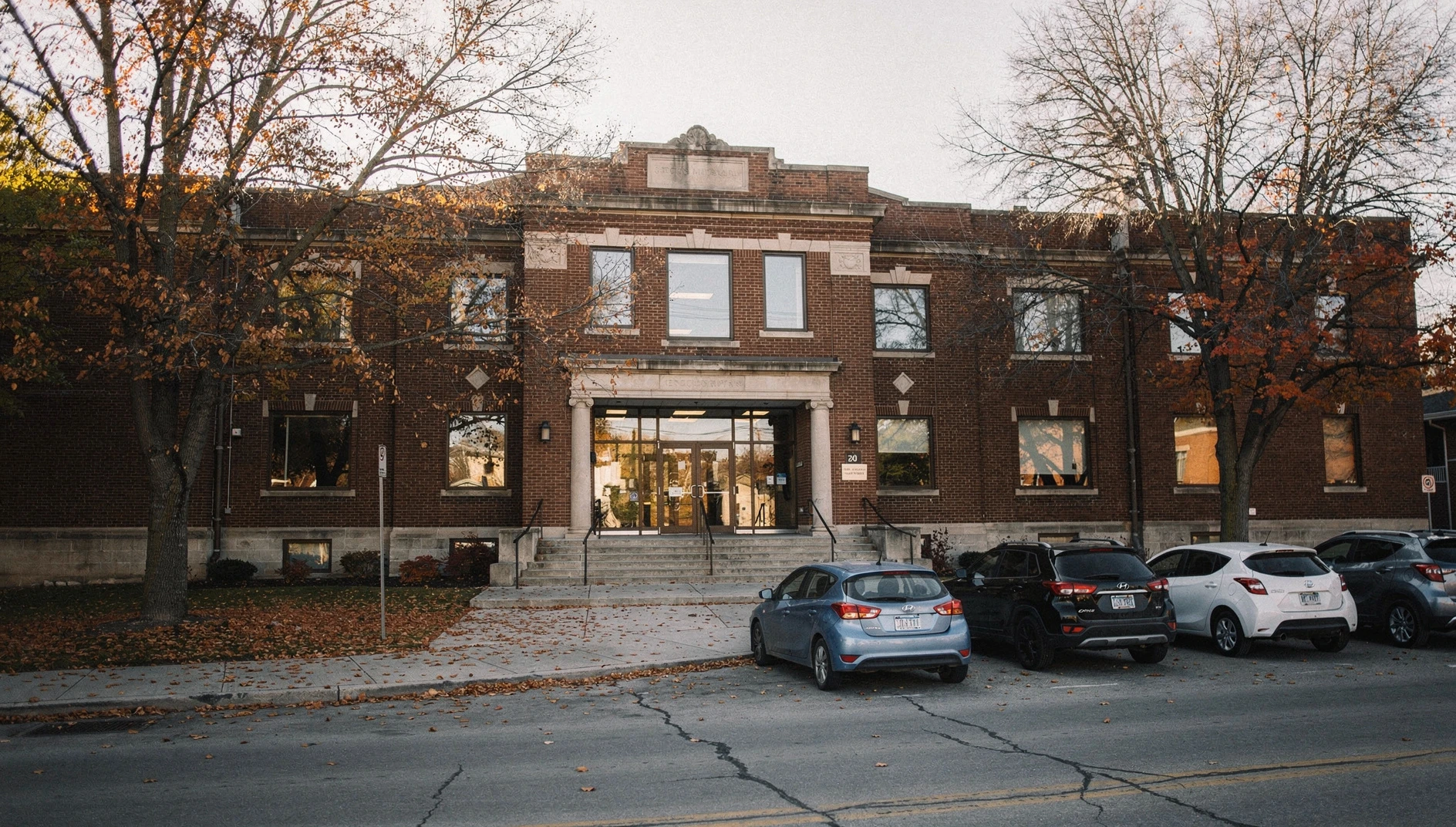 Red-brick municipal building exterior with autumn trees and fallen leaves on the sidewalk.