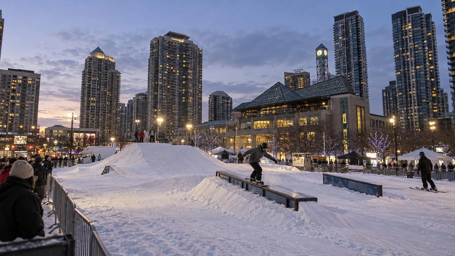 An outdoor urban ski park with ramps and rails set up in a snowy city square at dusk, with city buildings in the background.