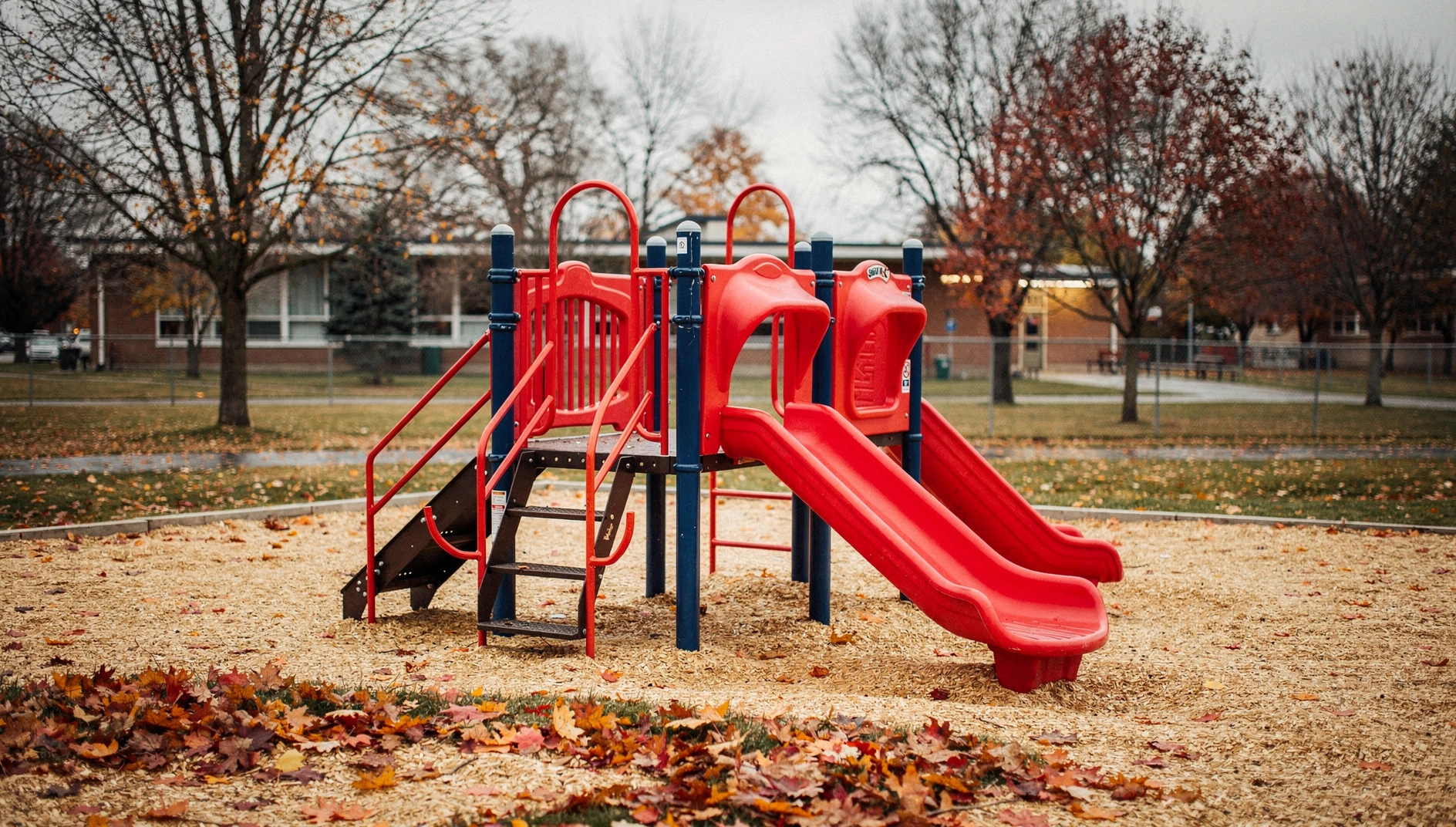 A new school playground with climbing equipment and slides surrounded by fallen autumn leaves.