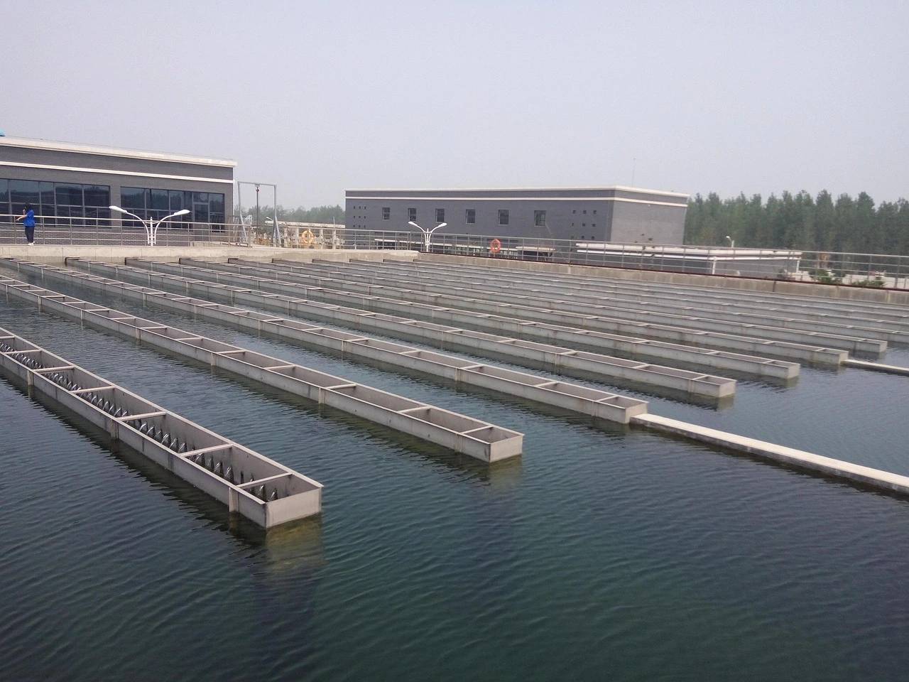An architectural view of a municipal water treatment facility with large industrial pipes and concrete structures under an autumn sky.