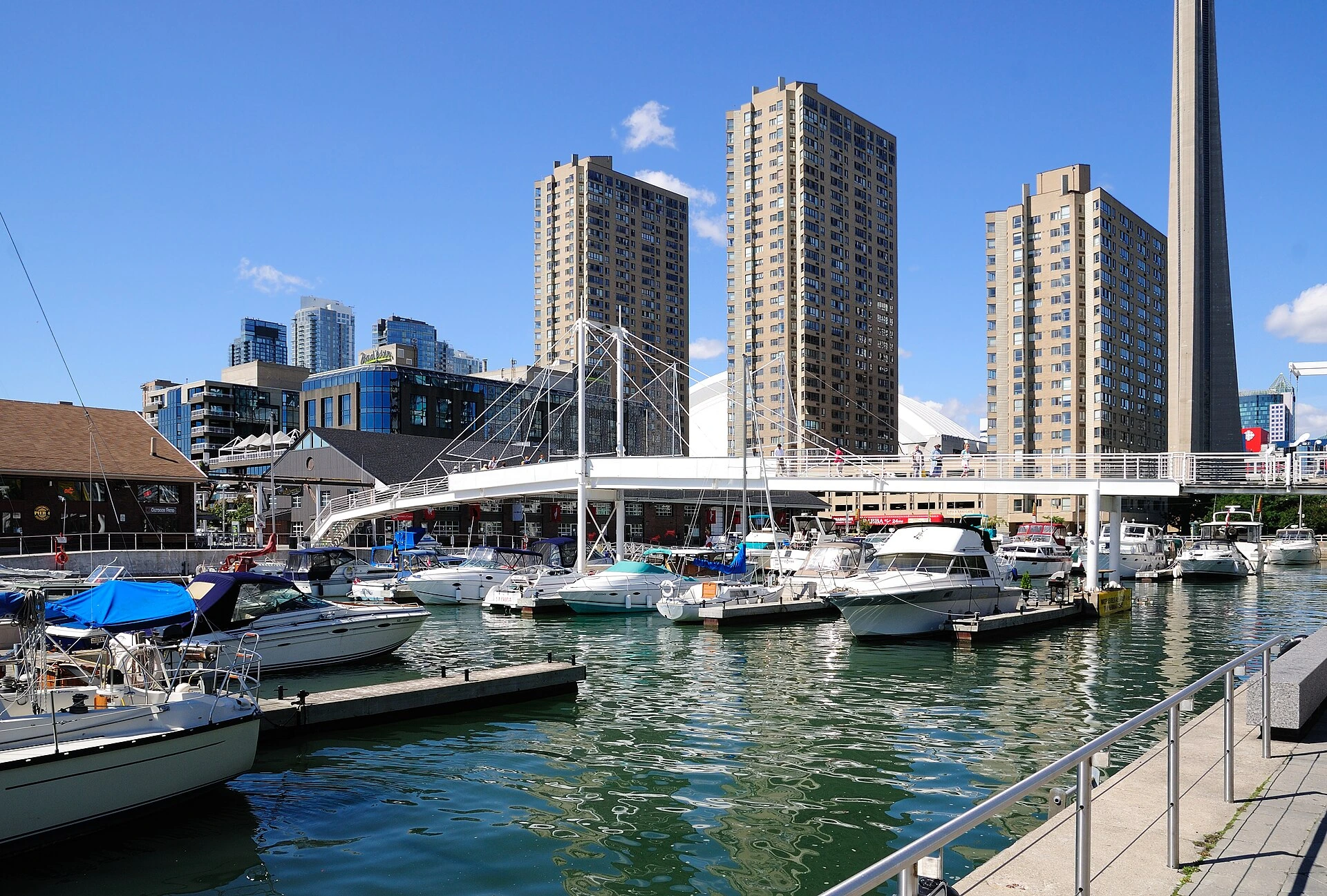 A steel cable pedestrian footbridge over a waterfront slip with the Toronto skyline behind it