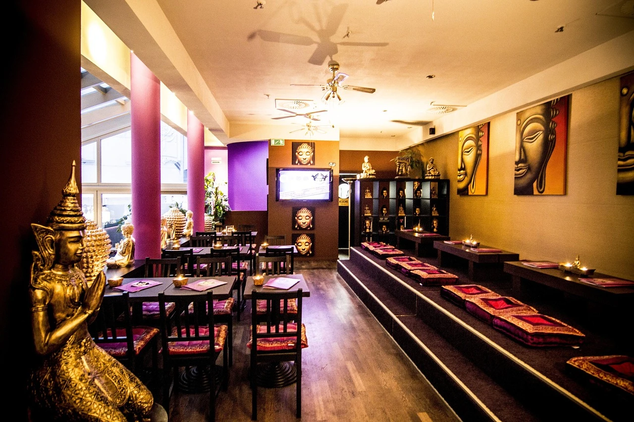 A warmly lit restaurant interior with wooden tables and rattan chairs beside a glass wall overlooking a busy shopping centre concourse.