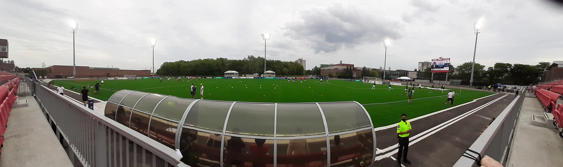 A women's soccer match in progress at a small outdoor stadium in Toronto, with a goalkeeper and players on the field in evening light.