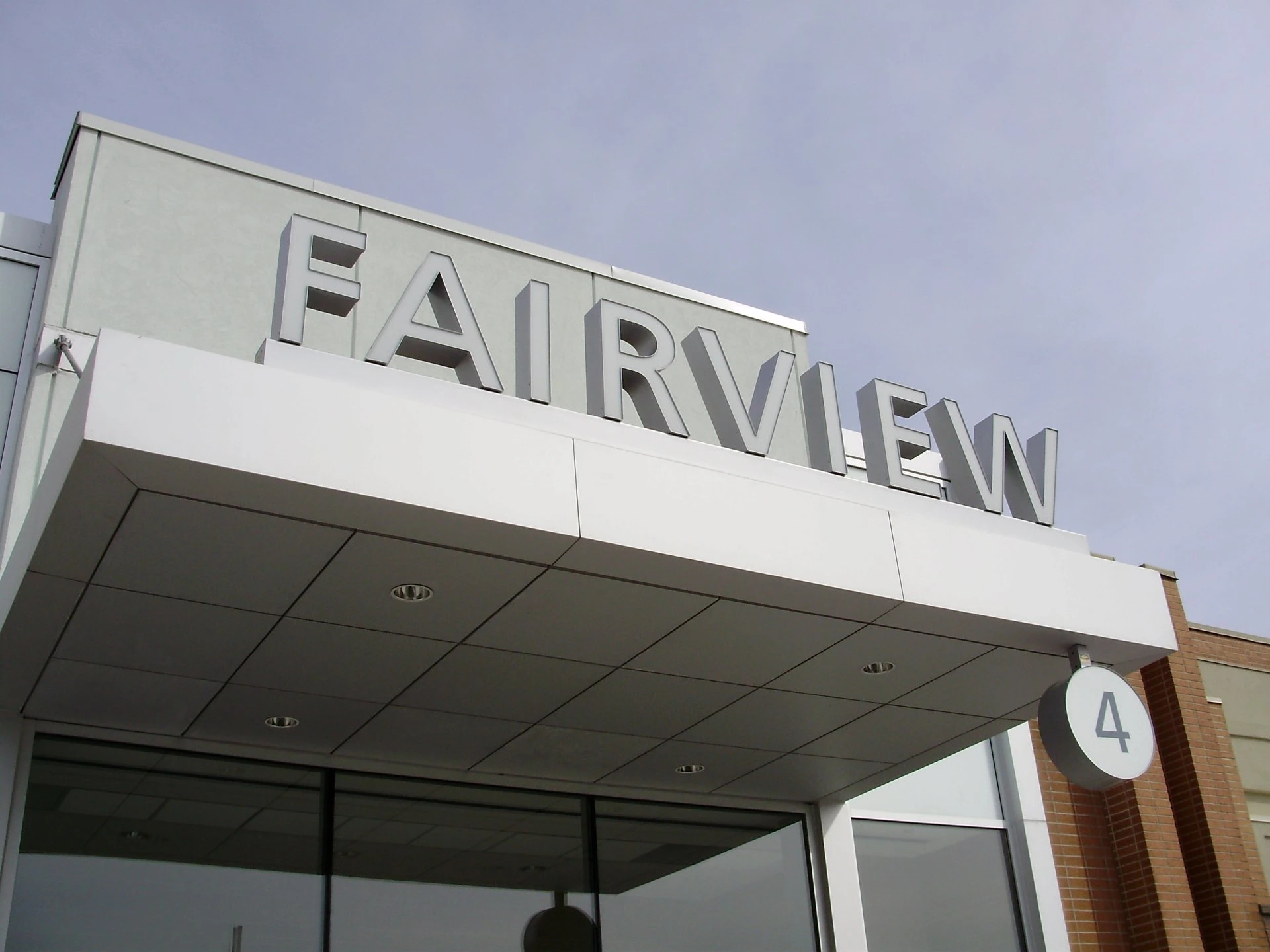 A construction crane above the concrete frame of a mid-rise apartment building under construction, with mature trees and apartment towers in the background on a sunny summer day.