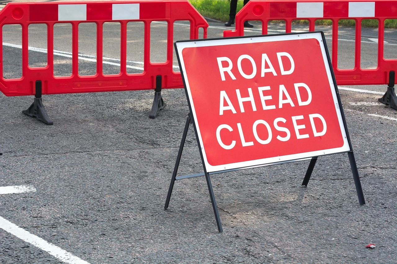 An orange road closure and detour sign on a leaf-covered suburban street, illustrative of the temporary closure in Pelham.