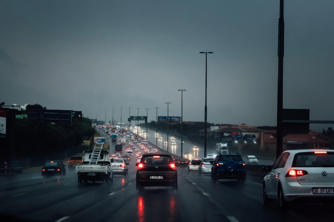 Cars with headlights on travel along a wet multi-lane highway in the rain under a grey summer sky.