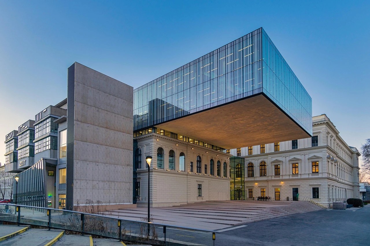 A modern, low-rise institutional building with large windows and greenery under a clear summer sky.