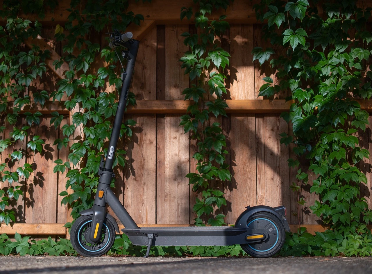 An electric kick scooter parked upright on a sidewalk beside a suburban plaza with autumn trees.
