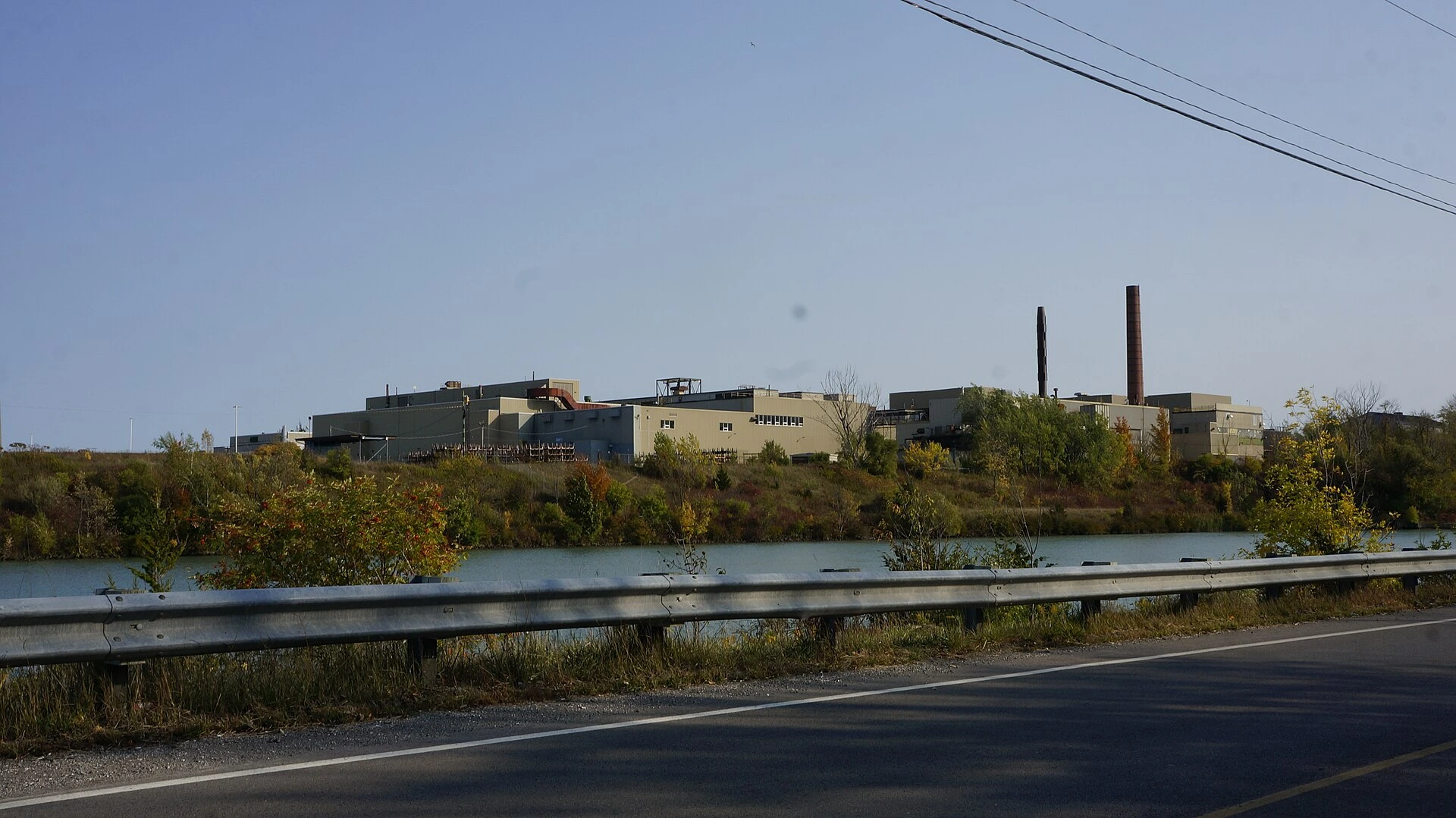 Abandoned brick industrial buildings behind a chain-link fence, surrounded by overgrown grass and rubble on a summer day