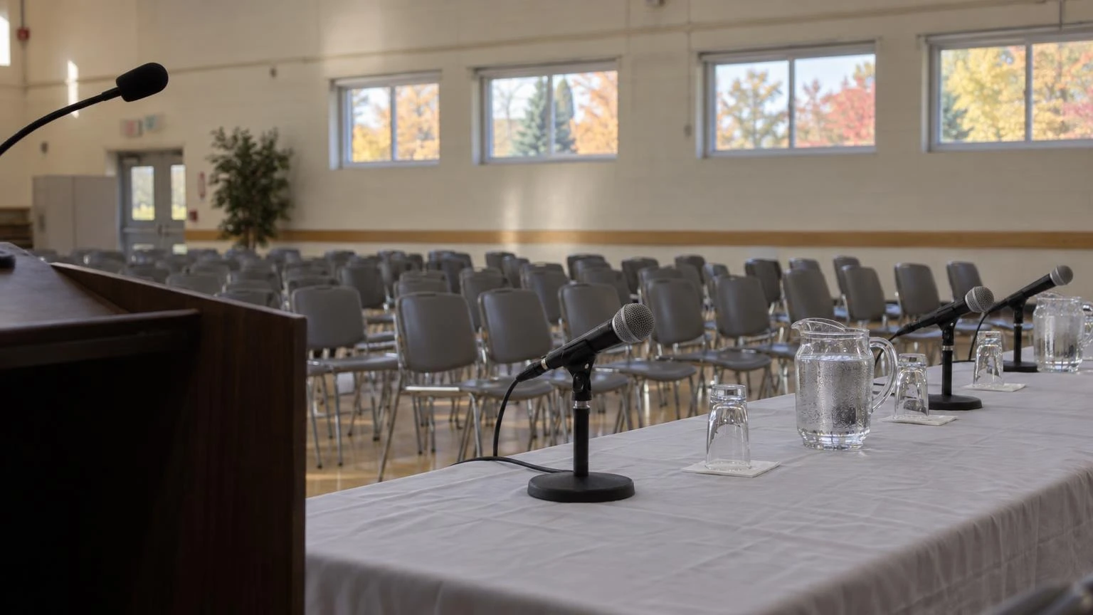 A close-up of a microphone and water glasses on a head table inside a community hall set up for a candidates' forum.