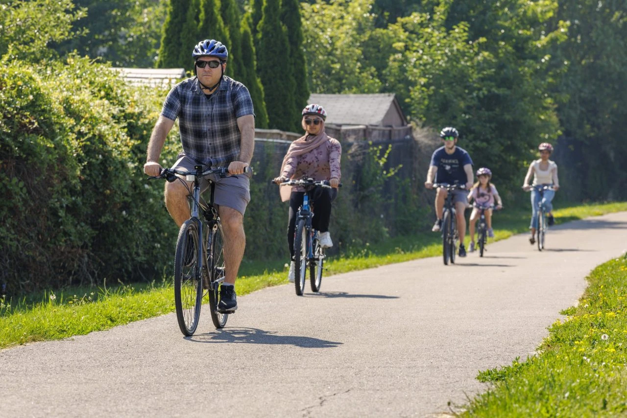 A helmeted cyclist rides along a tree-lined path with autumn foliage in a suburban park.