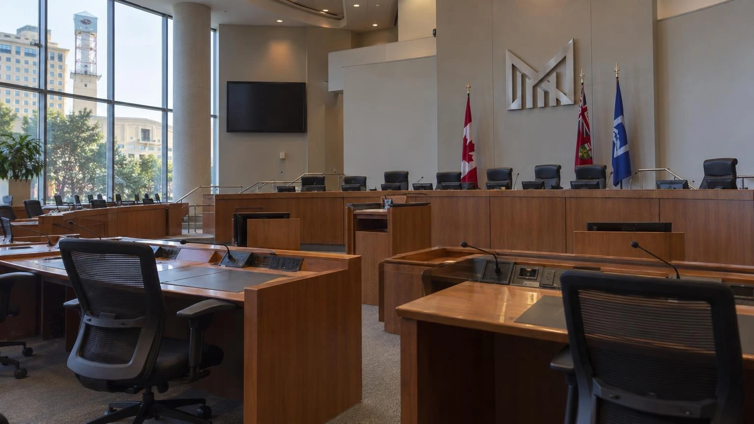 An empty Mississauga City Council chamber with wooden desks and microphones under soft indoor lighting.