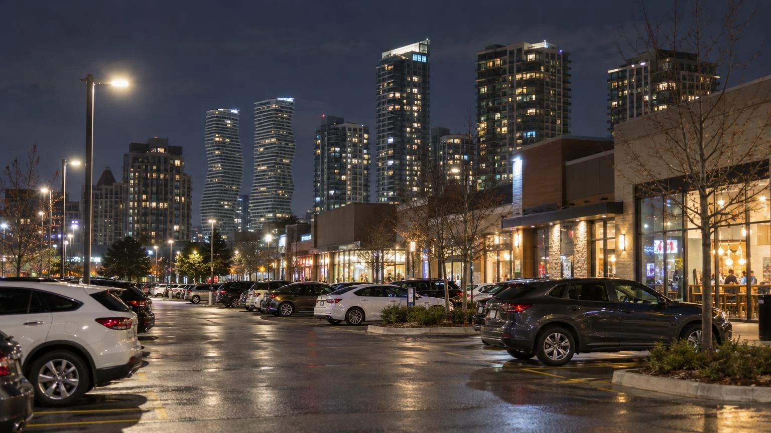 A street-level view of a commercial plaza in Mississauga at night with glowing storefronts and parked cars.