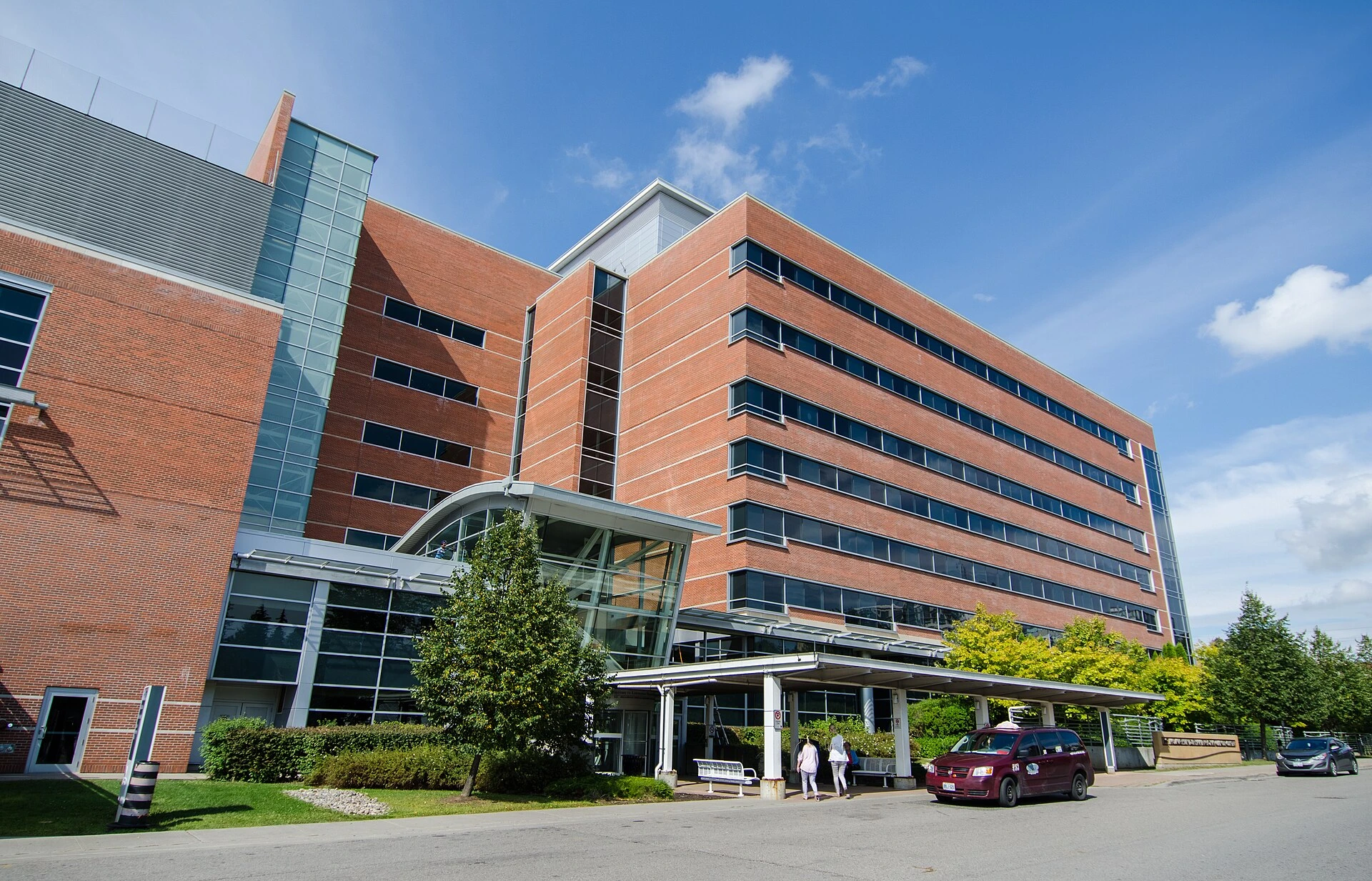 The exterior entrance of a community hospital with a covered drop-off area and green trees on an overcast day.