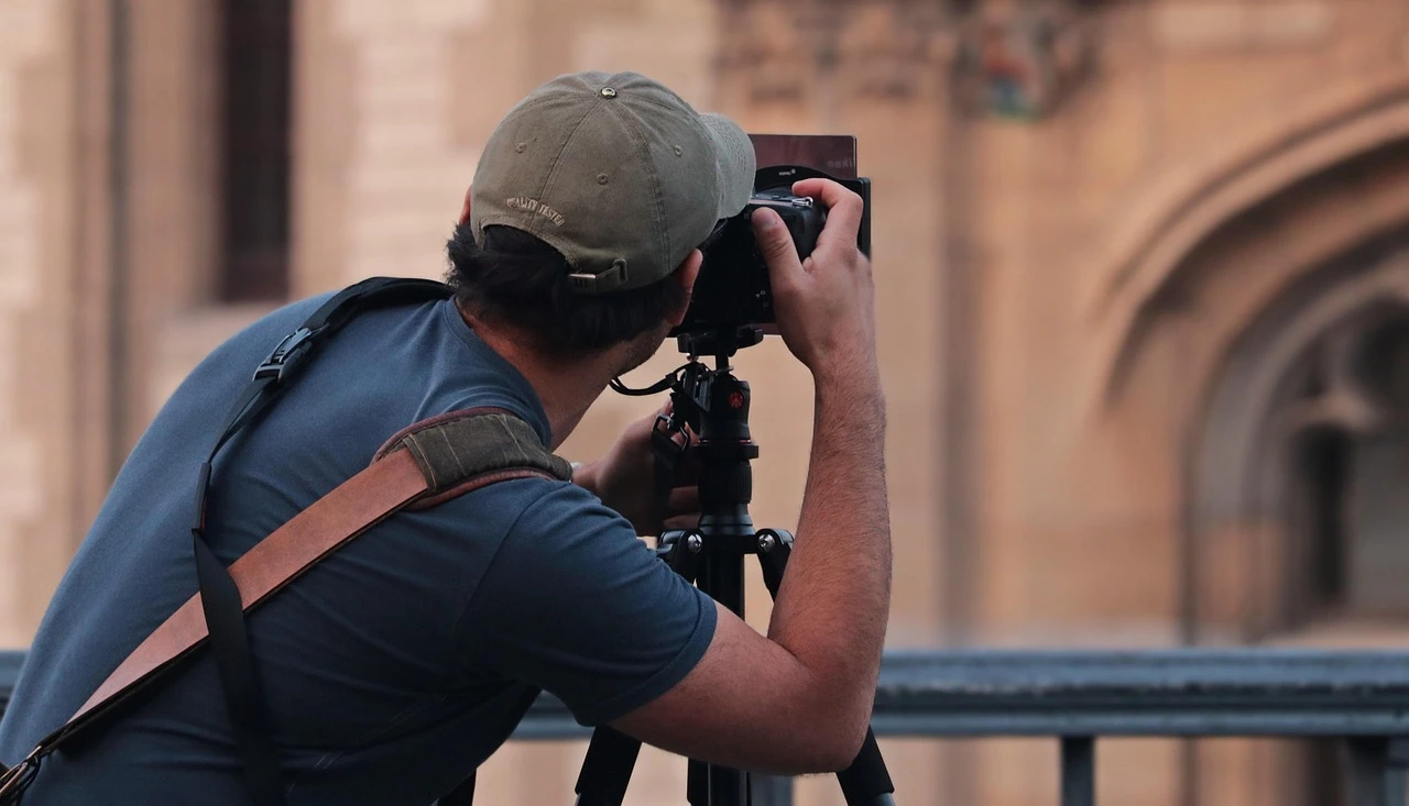 A photographer with a camera walks along a sunny downtown street lined with trees and storefronts.