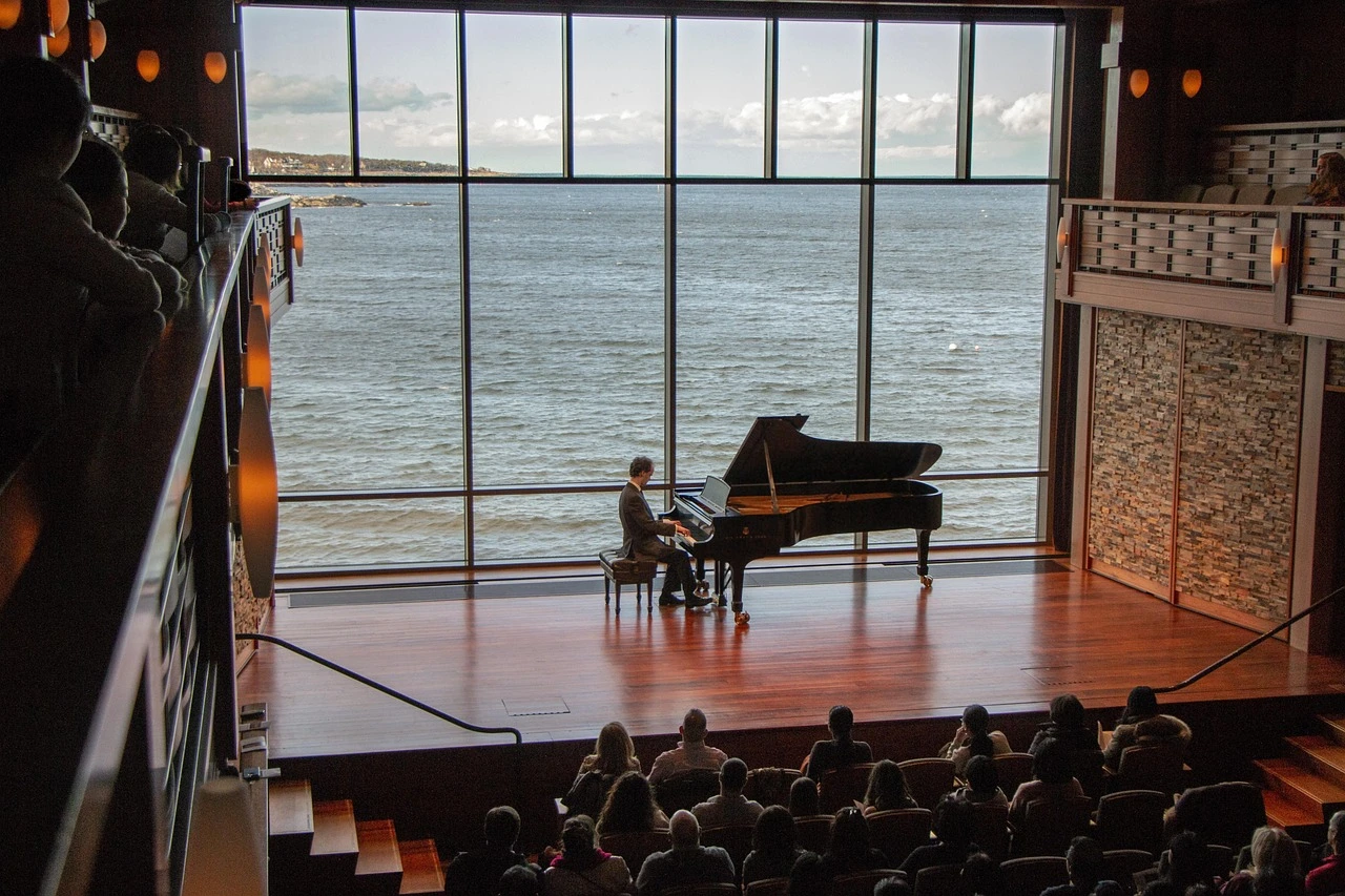 A grand piano on a small community hall stage with rows of empty chairs, winter light through tall windows.