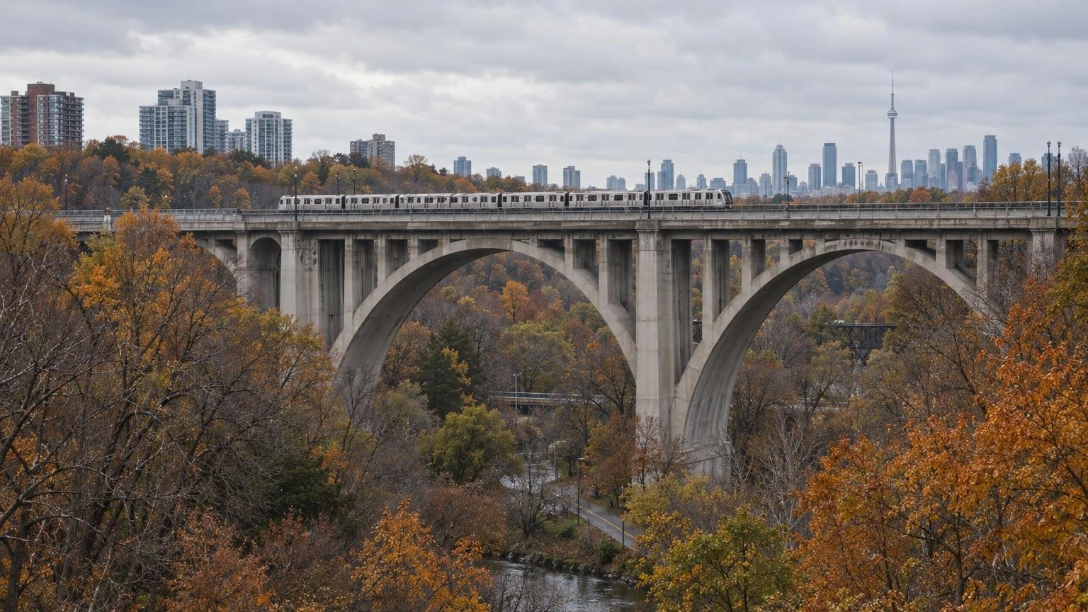 A concrete arch viaduct crossing a tree-lined valley with a subway train passing over the top deck in autumn.