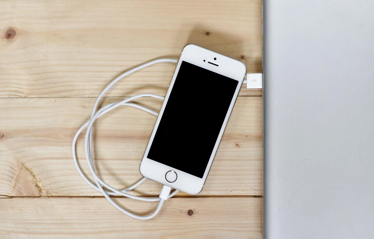 A phone and laptop connected to charging cables on a desk beside a window.
