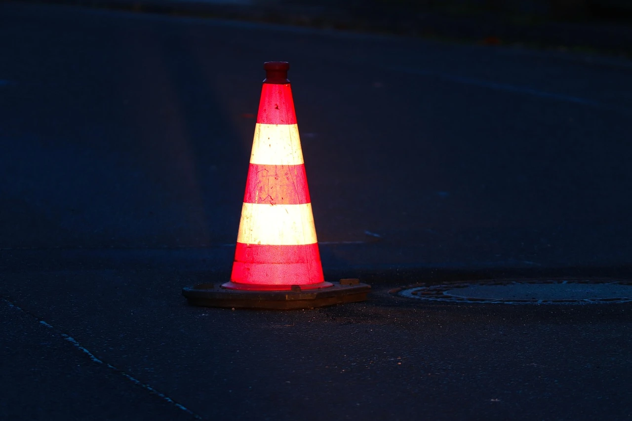 A construction zone on a rural two-lane road with orange pylons and a detour sign under autumn trees.